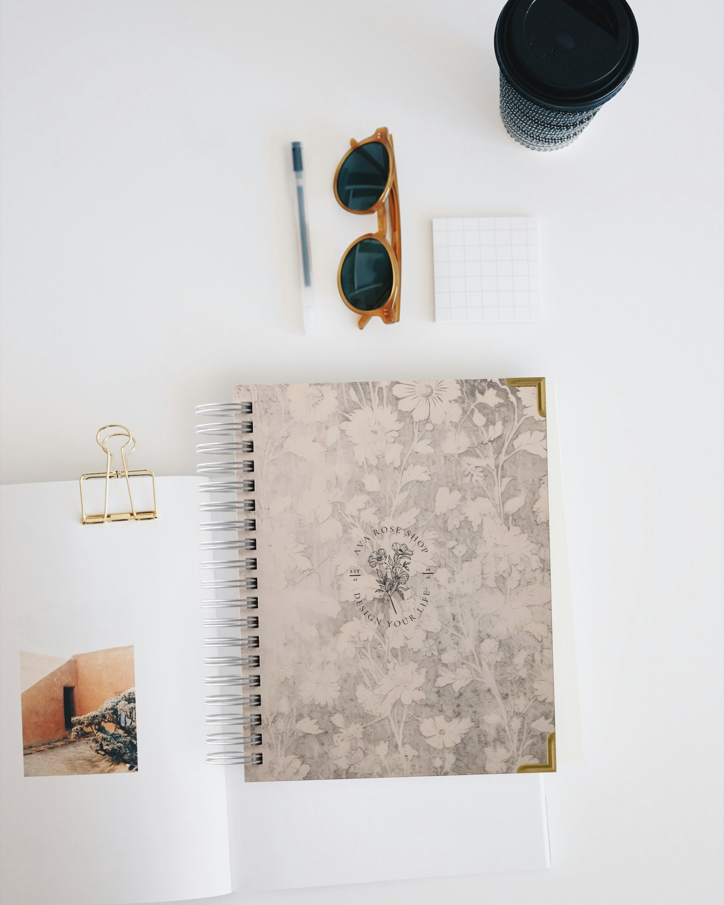 Planner with floral design, sunglasses, pen, and cup on a white surface