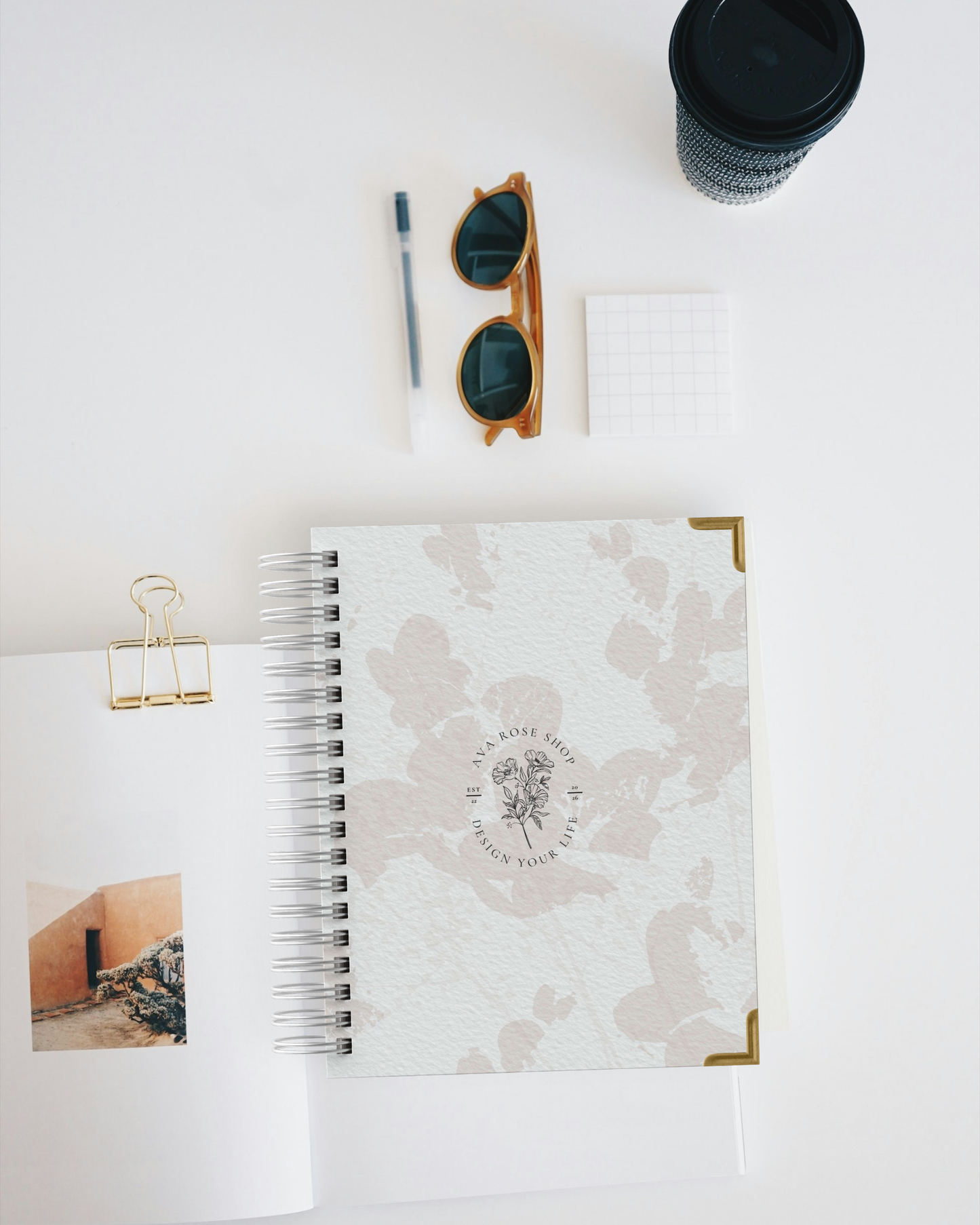 Spiral-bound planner with floral design, sunglasses, pen, and cup on a white surface