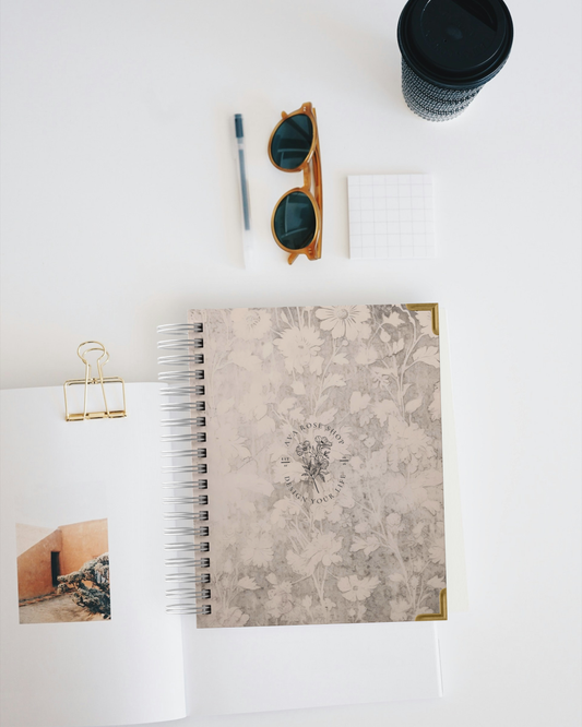 Planner with floral design, sunglasses, pen, and cup on a white surface