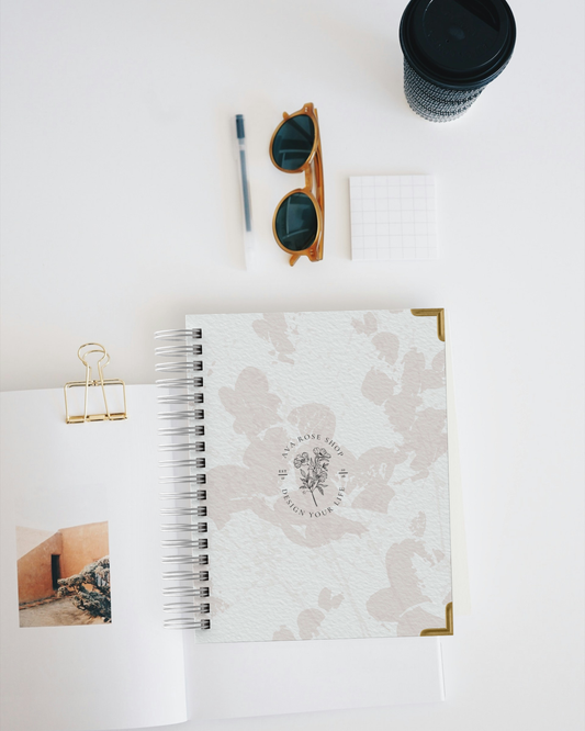 Spiral-bound planner with floral design, sunglasses, pen, and cup on a white surface