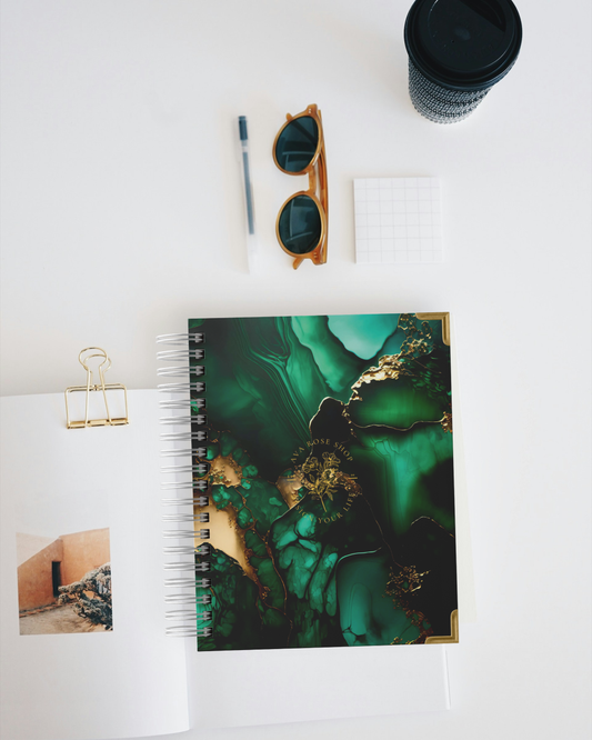 Planner with green and gold abstract design, sunglasses, pen, and coffee cup on a white surface